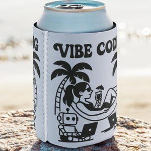 Peut inclure: Un refroidisseur de canette blanc avec le texte "VIBE CODING" et une illustration noire d'une personne se relaxant sur une plage avec un ordinateur portable, une boisson et un robot. Le refroidisseur de canette est sur un rocher près de la mer.