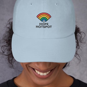 Puede incluir: Gorra de béisbol azul claro con visera curva. La gorra presenta un arcoíris y las palabras "HOPE HOTSPOT" en negro. El sombrero está hecho de un material suave y está diseñado para ser usado.