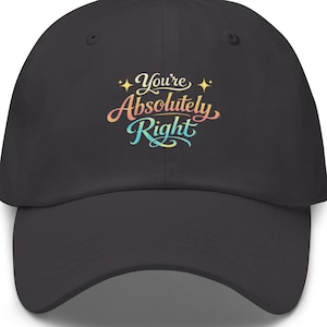 Puede incluir: Gorra de béisbol gris oscuro con visera curva. La gorra presenta la frase "You're Absolutely Right" en letras coloridas y estilizadas. El texto es en tonos de amarillo, rosa, azul y verde, con acentos de estrellas.