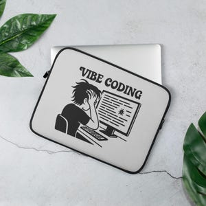 Puede incluir: Una funda para portátil gris claro con cremallera negra y un gráfico en blanco y negro. El gráfico representa a una persona con la cabeza entre las manos frente a una pantalla de ordenador con el texto "VIBE CODING".