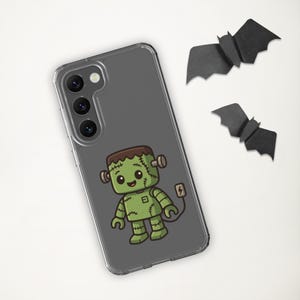 May include: A clear phone case featuring a cartoon Frankenstein's monster in green, brown, and black. The design includes stitches, bolts, and a power plug. Two black paper bats are placed near the phone case, creating a Halloween theme.