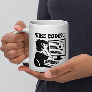 Puede incluir: Taza de cerámica blanca con la frase "Vibe Coding" y un gráfico negro de una persona estresada frente a una pantalla de ordenador con un icono de error. La taza está sujeta por una persona.