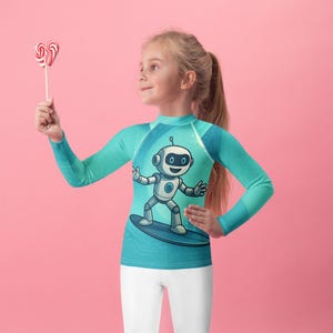May include: A turquoise long-sleeve shirt with a cartoon robot surfing on a blue surfboard. The robot has a white body, black eyes, and a smiling face. The shirt is paired with white leggings. A heart-shaped lollipop is held in the hand.