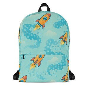 Peut inclure: Un sac à dos bleu clair avec un motif répété de fusées orange, de nuages blancs et de code binaire. Le sac à dos a des bretelles noires et une poignée supérieure noire. Les fusées ont des flammes jaunes et des détails bleus et orange.