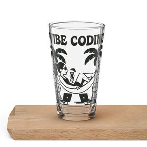 Pode incluir: Copo transparente com as palavras "VIBE CODING" em preto, apresentando uma ilustração a preto e branco de uma pessoa relaxando numa rede com um cocktail, palmeiras e laptops. O copo está sobre uma superfície de madeira.