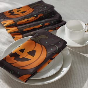 May include: A stack of dark gray Halloween napkins featuring orange jack-o'-lantern faces. The napkins are folded and placed on white plates, with a white teacup and saucer in the background. The design includes a pumpkin, swirls, and small green dots.