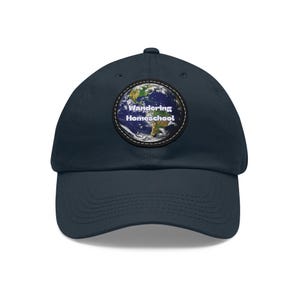 Puede incluir: Gorra de béisbol azul marino con un parche circular con un diseño de globo terráqueo. El parche tiene las palabras "Wandering Homeschool" en blanco. La gorra está hecha de un material suave y tiene una visera curva.