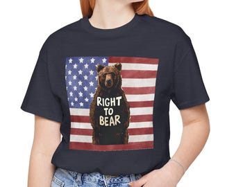 Puede incluir: Camiseta azul marino con una ilustración de un oso pardo y el texto "RIGHT TO BEAR" superpuesto a un diseño de bandera estadounidense descolorida. La camiseta está hecha de un material suave y cómodo.