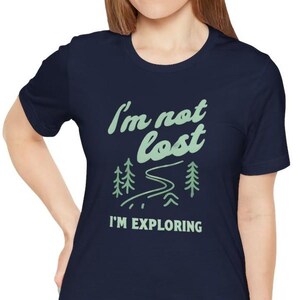 May include: Navy blue t-shirt with the text "I'm not lost, I'm exploring" in a light green color. The design includes a winding road and two trees. The shirt is a crew neck.