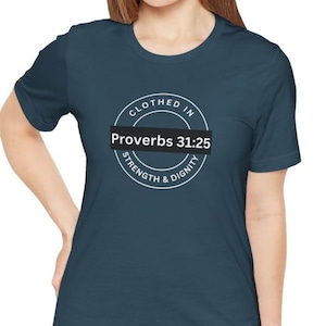 Puede incluir: Camiseta de color verde azulado con el texto "CLOTHED IN STRENGTH & DIGNITY" en un diseño circular. Las palabras "Proverbios 31:25" están en un rectángulo negro. La camiseta es de material suave.