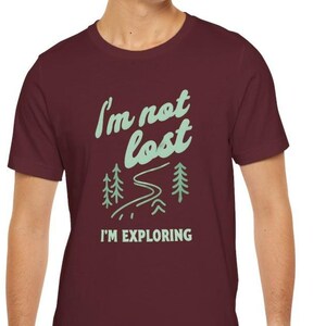 May include: A maroon t-shirt with the text "I'm not lost, I'm exploring" in a light green color. The design includes stylized trees and a winding path, evoking a sense of adventure and outdoor exploration.