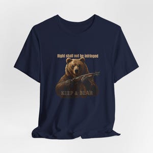 Peut inclure: T-shirt bleu marine avec un ours brun tenant un fusil. Le texte "Right shall not be infringed" est au-dessus de l'ours, et "KEEP & BEAR" est en dessous. Le motif est centré sur le t-shirt.