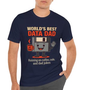 Könnte beinhalten: Marineblaues T-Shirt mit dem Text "WORLD'S BEST DATA DAD" in Weiß und Rot. Eine Cartoon-Diskettenfigur hält eine Kaffeetasse mit der Aufschrift "#1 DAD". Der Text "Running on coffee, code, and dad jokes" steht darunter.