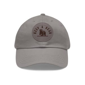 Puede incluir: Gorra de béisbol gris con visera curva. La gorra presenta un parche circular con las palabras "KEEP & BEAR" y un gráfico de oso marrón. El sombrero está hecho de un material suave y tiene una correa ajustable.
