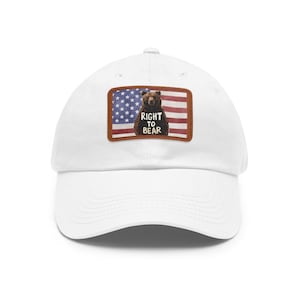 Puede incluir: Gorra de béisbol blanca con un parche que presenta un diseño de la bandera estadounidense y un oso pardo. El texto "RIGHT TO BEAR" está impreso en el parche. La gorra tiene una visera curva y un diseño clásico de seis paneles.