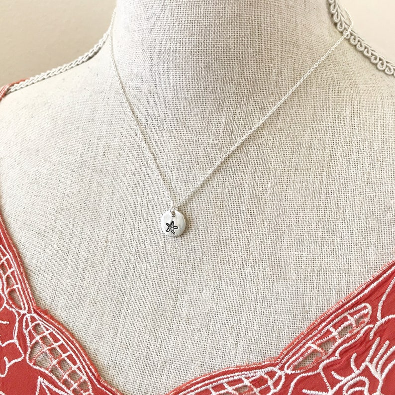 May include: A silver necklace with a small, round pendant featuring a starfish design. The necklace is displayed on a white mannequin with a red and white patterned top.