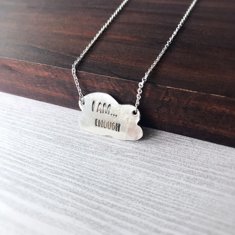 May include: A silver necklace with a cloud-shaped pendant that reads "I AM... ENOUGH".