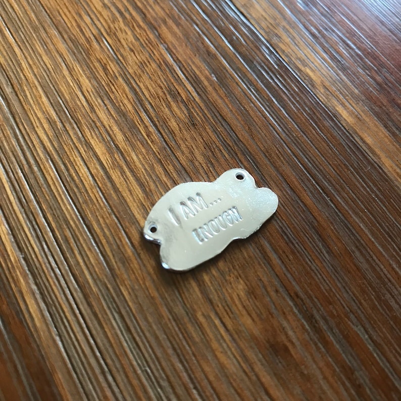 May include: Silver charm with a cloud shape and the text "I am... enough" engraved on it.