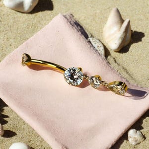 Puede incluir: Un piercing para el ombligo de color dorado con un diseño de barra curva. El anillo presenta una gema grande, transparente y de talla redonda en el centro, con dos gemas más pequeñas y una gema rectangular en el extremo.