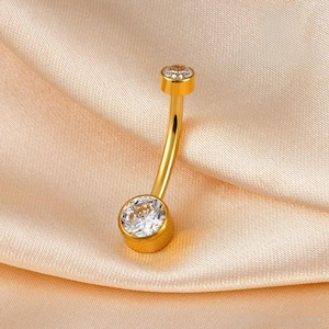 May include: A gold-colored belly button ring with a curved barbell design. The ring features a large, clear, round-cut gemstone at the bottom and a smaller gemstone at the top. The jewelry is set against a peach-colored fabric.