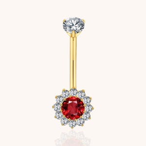May include: A gold-colored belly button ring with a clear, round gemstone at the top and a cluster of clear gemstones surrounding a red gemstone at the bottom. The jewelry is set against a soft, neutral background.