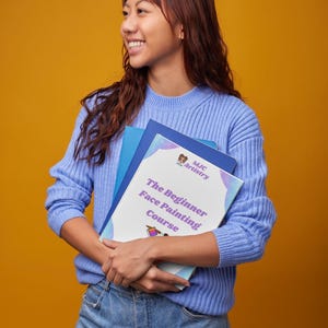 May include: A person in a light blue sweater and jeans holds a book titled "The Beginner Face Painting Course." The book has a white cover with colourful accents and the MJC Artistry logo. The background is a solid orange colour.