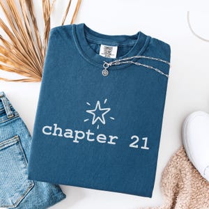 Peut inclure: Un t-shirt bleu Comfort Colors avec le texte "chapter 21" et une étoile. Un collier argenté avec un pendentif soleil est posé sur le t-shirt. Le t-shirt est plié avec un jean et une chaussure blanche.