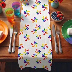 May include: A white table runner with a repeating pattern of colorful, swirling designs. The runner is set on a wooden table, alongside plates, silverware, and glasses. The table is set for a meal.