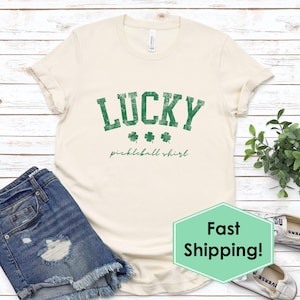 May include: Cream-colored t-shirt with the word "LUCKY" in distressed green lettering, along with three green shamrocks. Below the word "LUCKY" is the text "pickleball shirt" in a script font. The shirt is paired with denim shorts.