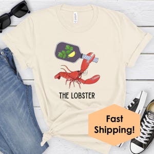 May include: Cream-colored t-shirt featuring a red lobster holding a bottle with parsley and a lemon slice. The text "THE LOBSTER" is printed below the lobster graphic. The shirt is paired with blue jeans and black sunglasses.