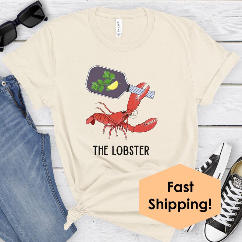 May include: Cream-colored t-shirt featuring a red lobster holding a bottle with parsley and a lemon slice. The text "THE LOBSTER" is printed below the lobster graphic. The shirt is paired with blue jeans and black sunglasses.