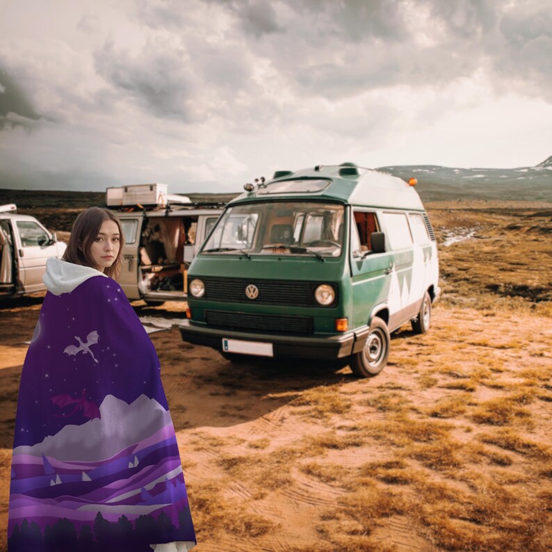 May include: A hooded cloak with a purple and white design featuring a dragon, mountains, and clouds. A green and white van is parked in a field under a cloudy sky.