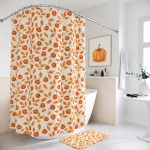 May include: A shower curtain featuring a repeating pattern of orange pumpkins and golden-brown autumn leaves on a cream-colored background. The fabric hangs from a curved metal rod, and a matching rectangular bath mat lies on the floor below.