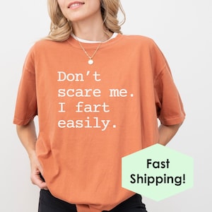 May include: An orange t-shirt with the text "Don't scare me. I fart easily." in white. The shirt has a relaxed fit and a round neckline. The shirt is a solid color and is made of a soft material.