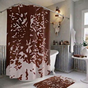 Puede incluir: Una cortina de ducha con un patrón de piel de vaca marrón y blanco colgada de una barra de metal curva. La cortina está colocada alrededor de una bañera blanca. Una alfombrilla de baño rectangular a juego yace en el suelo frente a la bañera.