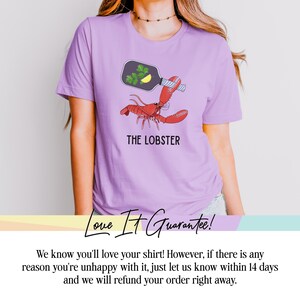 May include: A light purple t-shirt featuring a red lobster illustration with a bottle and the text "THE LOBSTER". The bottle contains parsley and a lemon slice. The shirt is made of a soft material.