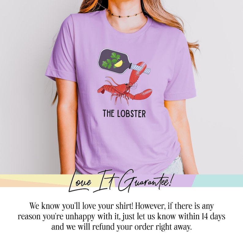 May include: A light purple t-shirt featuring a red lobster illustration with a bottle and the text "THE LOBSTER". The bottle contains parsley and a lemon slice. The shirt is made of a soft material.
