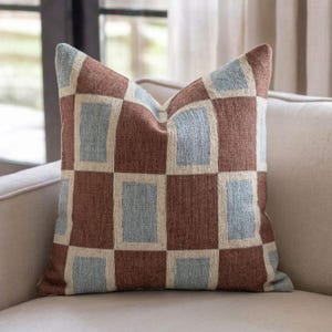May include: A square throw pillow featuring a geometric pattern of alternating brown and light blue rectangular blocks separated by thin cream-colored lines. The fabric has a visible woven texture throughout the entire surface of the cushion.