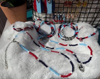 May include: A collection of handmade jewelry, including necklaces, bracelets, and earrings. The pieces feature red, white, and blue beads, with football-themed charms. The jewelry is displayed on a fluffy white surface.