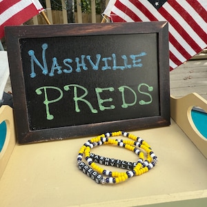 May include: A chalkboard sign with the words "Nashville Preds" in blue and green chalk sits on a tray with two beaded bracelets. The bracelets are yellow, white, and blue, spelling out "Nashville Preds". Two American flags are in the background.