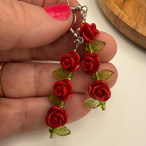 May include: A pair of dangling earrings featuring red rose-shaped beads with green leaf accents. The earrings are suspended from silver-colored hooks. The roses are a vibrant red, and the leaves are a translucent green.