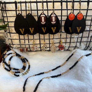 May include: A collection of earrings and beaded necklaces. The earrings include black teardrop shapes with a gold "V", football, basketball, and other sports-themed designs. The necklaces are black and gold beaded.