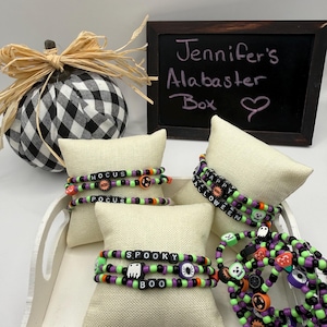 May include: A collection of Halloween-themed beaded bracelets featuring black, purple, and lime green plastic beads. The bracelets include letter beads spelling HOCUS POCUS, SPOOKY BOO, and HAPPY HALLOWEEN, along with charms shaped like ghosts, spiders, and pumpkins.