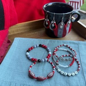 May include: A collection of Ohio State Buckeyes themed jewelry and accessories. Includes beaded bracelets, earrings, and a black mug. The bracelets feature red, gray, and white beads, with some spelling out "OHIO" and "UCKEYES". The earrings are teardrop-shaped.