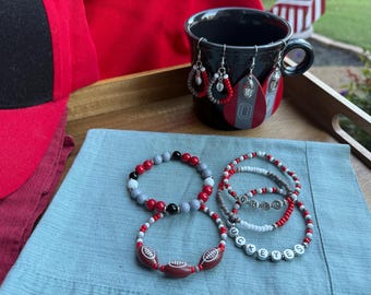 May include: A collection of Ohio State Buckeyes themed jewelry and accessories. Includes beaded bracelets, earrings, and a black mug. The bracelets feature red, gray, and white beads, with some spelling out "OHIO" and "UCKEYES". The earrings are teardrop-shaped.