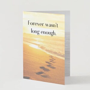 Puede incluir: Una tarjeta de felicitación plegada con una escena de playa. La tarjeta presenta una playa arenosa con huellas que conducen al océano bajo un cielo brillante y soleado. El texto "Forever wasn't long enough" está impreso en la parte delantera.