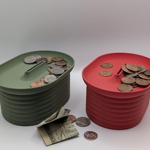 Op de afbeelding: Twee spaarpotten, één groene en één rode, elk gevuld met munten. De spaarpotten hebben een uniek ontwerp met een gleuf aan de bovenkant om geld in te doen. Een paar biljetten en munten liggen verspreid rond de groene spaarpot.