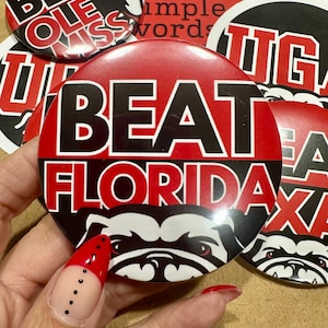 May include: A collection of red, black, and white round buttons with collegiate sports themes. One button reads "BEAT FLORIDA" with a bulldog graphic. Other buttons display team names and slogans.