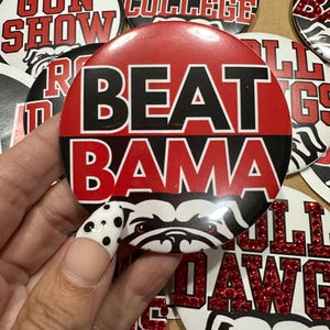 May include: A collection of round buttons with various text and graphic designs. One button reads "BEAT BAMA" in red and black. Other buttons say "I MISS COLLEGE", "Welcome to the GUN SHOW", and "BULLDAWG".