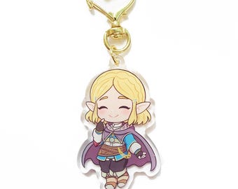 May include: A clear acrylic keychain featuring a cartoon character with blonde hair, wearing a blue and purple outfit. The keychain has a gold-colored heart-shaped clasp.